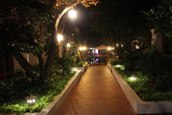 A nighttime pathway flanked by trees and illuminated by street lamps, leading to a building with a covered entrance.
