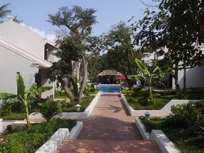 A pathway leading to a pool area with a thatched-roof structure, surrounded by lush greenery and white walls.
