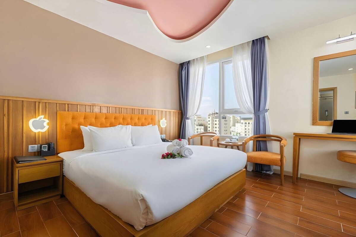 Une chambre d’hôtel avec un grand lit, une tête de lit en bois, un bureau avec une chaise et une vue sur la ville par la fenêtre.