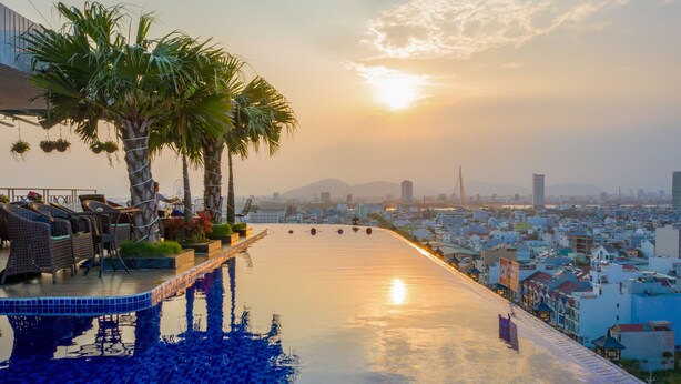 A rooftop infinity pool with lounge chairs and palm trees, overlooking a cityscape with buildings and a bridge. - Sea Queen Hotel (Da Nang)