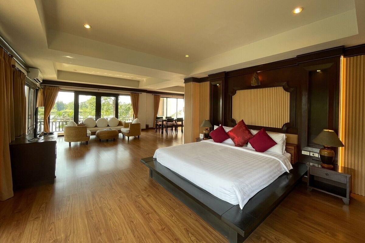 A spacious hotel room with a large bed, a seating area with chairs, and a view of the outdoors through large windows.