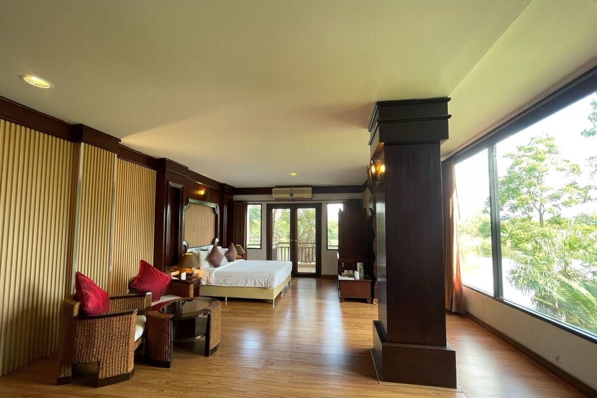 A spacious hotel room with a large bed, a seating area with wicker furniture, and a view of greenery through large windows.