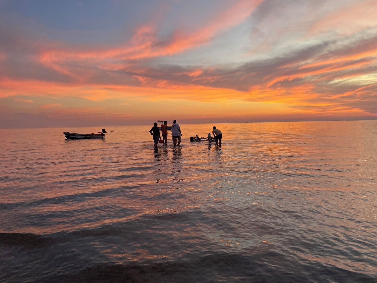 A sunset over a calm sea with a boat and people standing in the water.