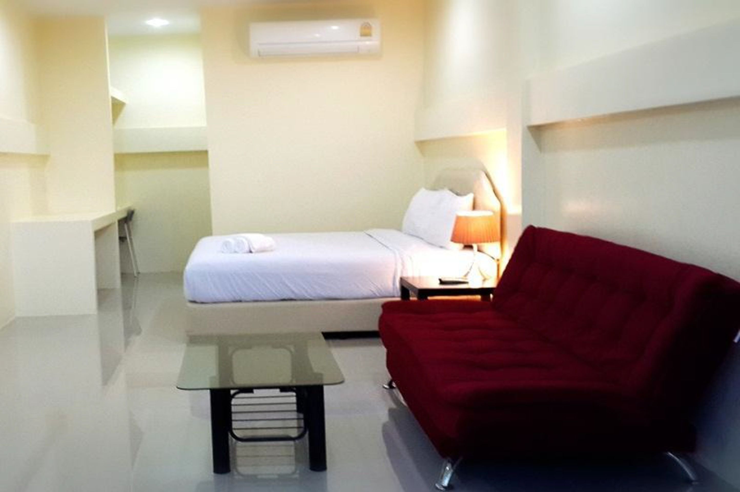 A small, modern hotel room with a bed, a red sofa, a small table, and an air conditioning unit on the wall.