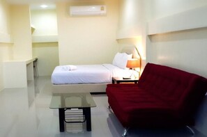 Une petite chambre d’hôtel moderne, équipée d’un lit, d’un canapé rouge, d’une petite table et d’un climatiseur fixé au mur.