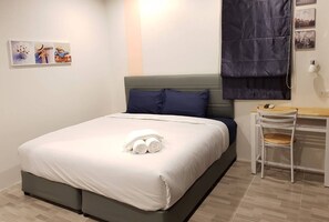 Un lit bien fait, avec une couette blanche et des serviettes pliées, une tête de lit grise, un bureau en bois avec une chaise et un téléviseur fixé au mur.