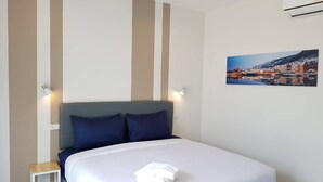 A neatly made bed with white linens and two pillows, a small bedside table, and a wall-mounted picture of a snowy town. - The Cube Resort (Chanthaburi)
