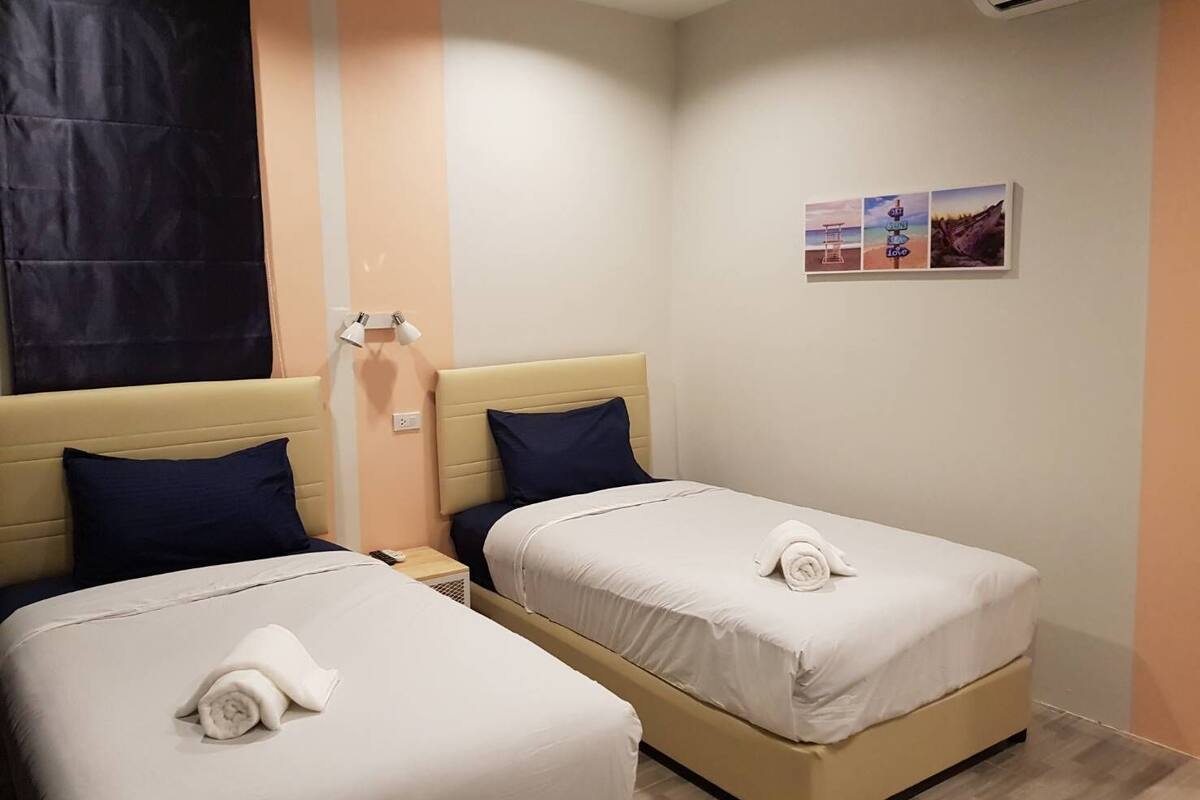 A hotel room with two beds, each with a pillow and a rolled towel. There is a nightstand with a lamp, and a wall-mounted air conditioning unit.
