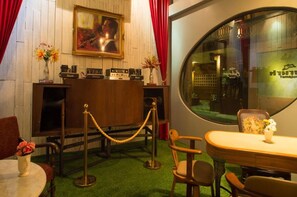 A room with a wooden reception desk, a painting on the wall, a round table with chairs, and a window with a view of the outside. - Tamajun Hotel (Chanthaburi)