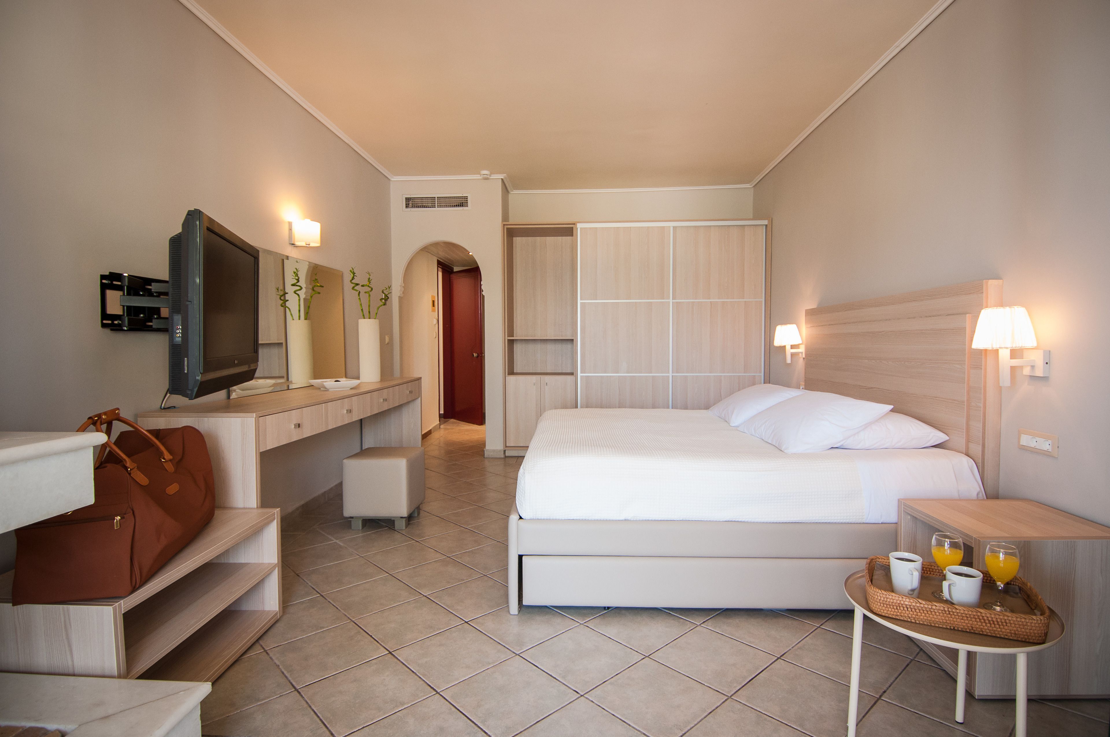 A hotel room with a bed, bedside table with a tray of breakfast, a desk with a TV, a wardrobe, and a bathroom visible through an archway.