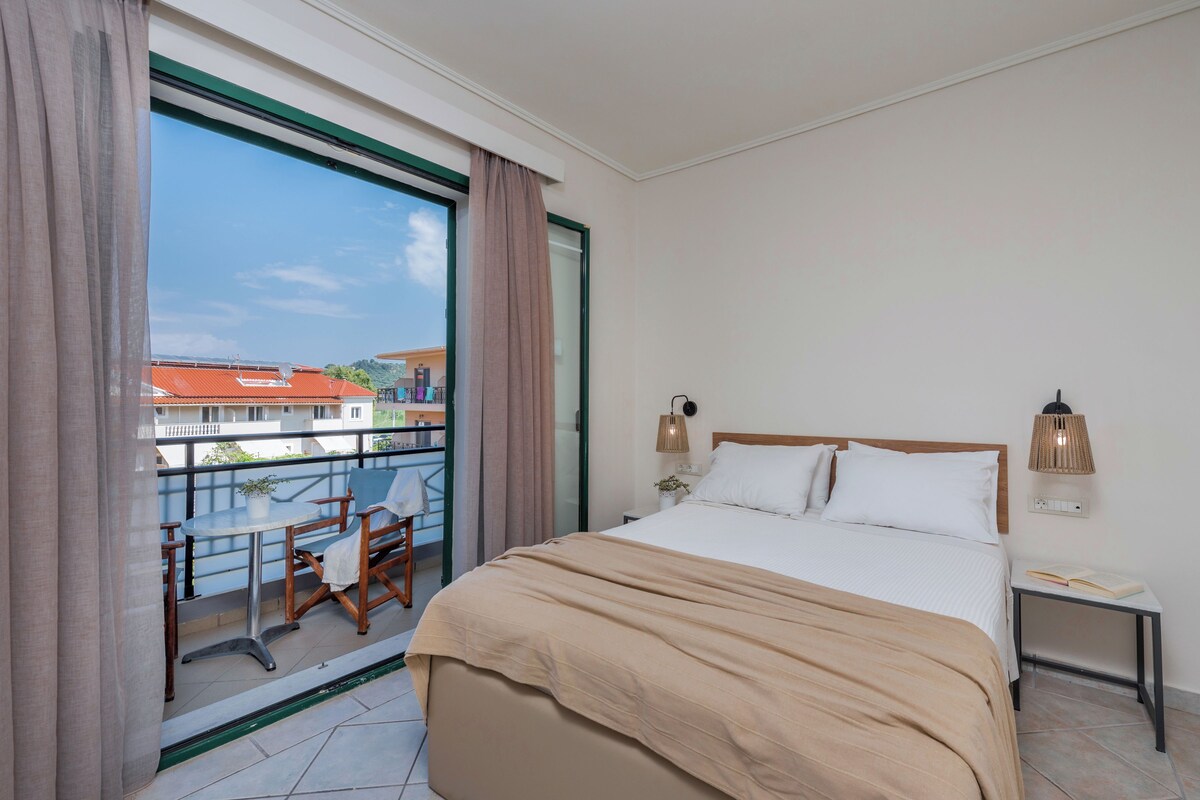 Hotelkamer met een bed, bedlampen, een klein tafeltje en uitzicht op gebouwen door een groot raam.