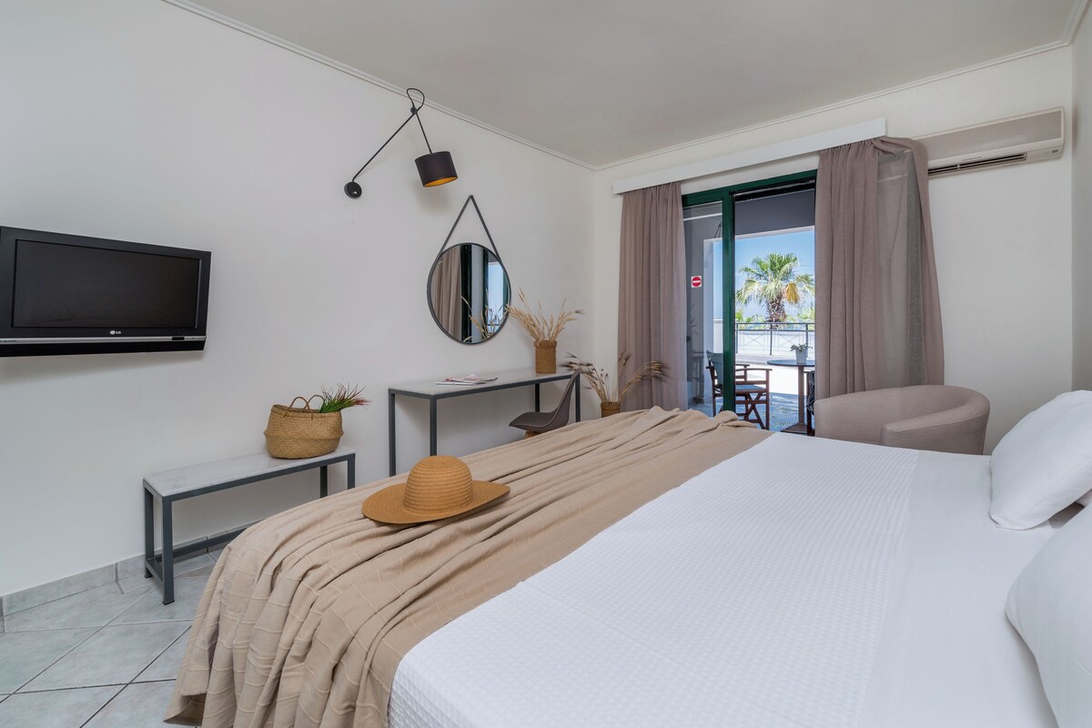 A hotel room with a bed, a TV, a desk, a chair, a balcony with a view of palm trees, and an air conditioning unit.