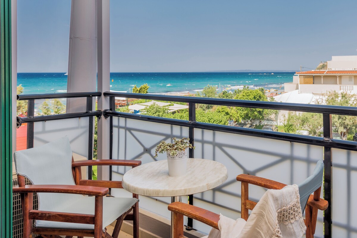 Een balkon met uitzicht op zee, houten stoelen, een klein rond tafeltje en een bloempot.