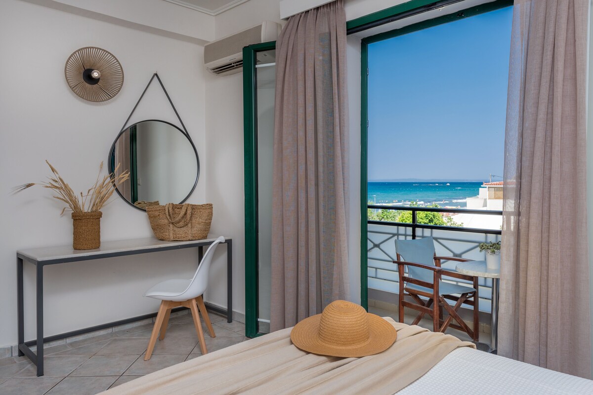 A bedroom with a bed, a desk, a chair, a large mirror, and a view of the sea through a balcony.
