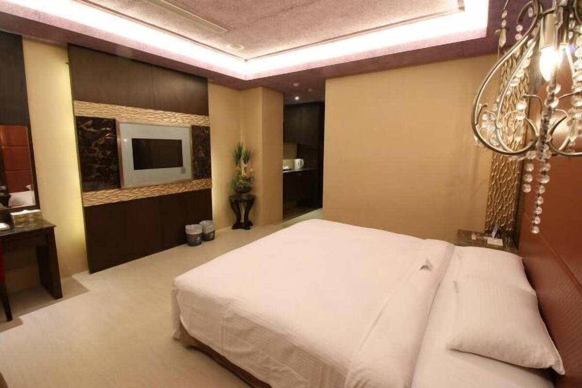 A hotel room with a large bed, a flat-screen TV mounted on the wall, a desk with a chair, and a decorative chandelier.