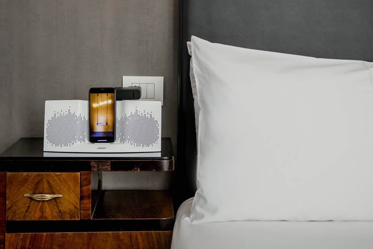 A neatly made bed with white sheets, a bedside table with a lamp, and a charging station with a smartphone on it.