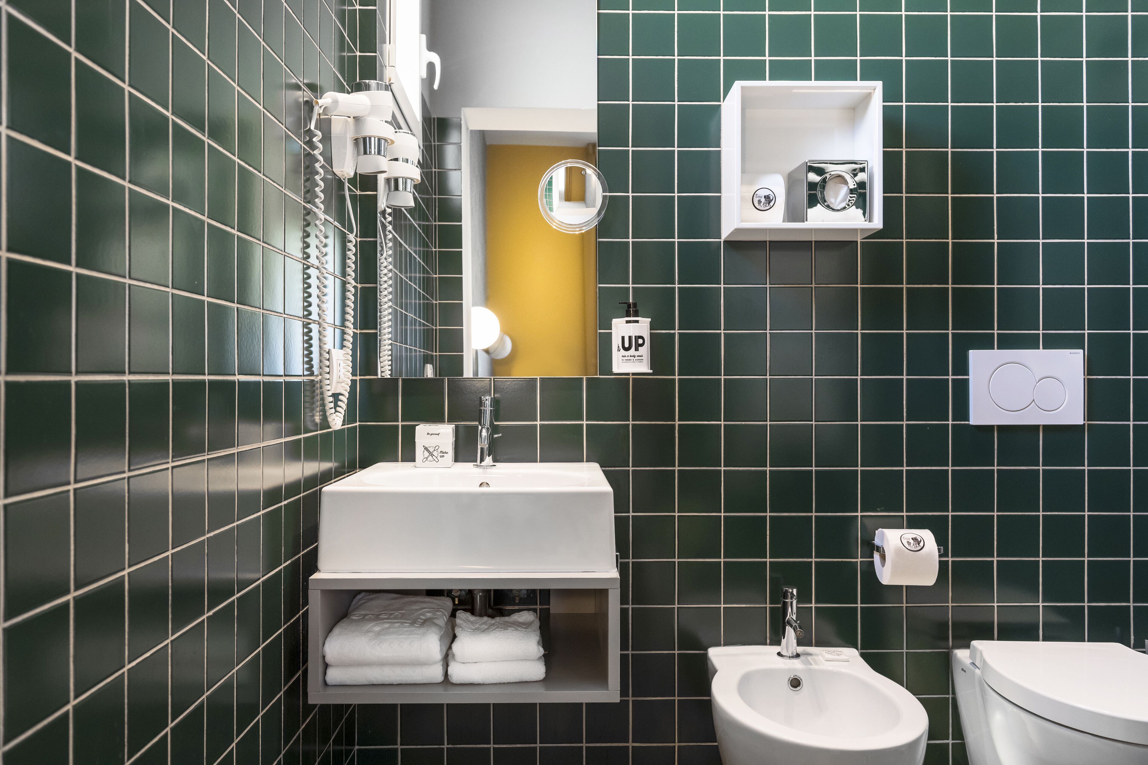 Un baño moderno con lavamanos blanco, un bidé y un inodoro. Las paredes están cubiertas con baldosas verdes y una pared de acento amarilla.