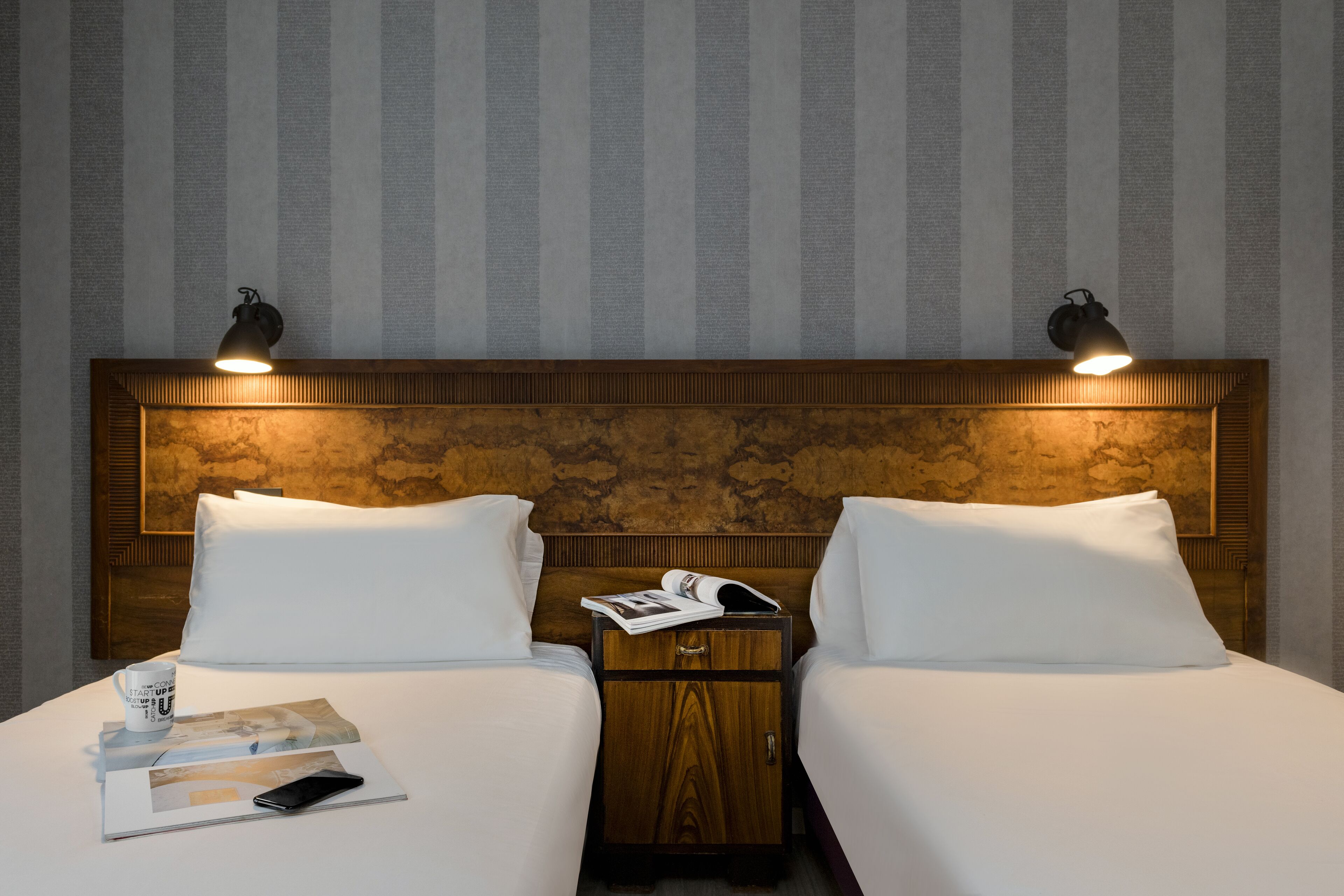 A double bed with a wooden headboard, two bedside tables with lamps, a magazine, and a smartphone on the bed.