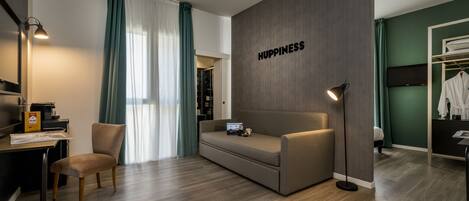 A modern hotel room with a sofa, a desk, a chair, a laptop, and a wardrobe.