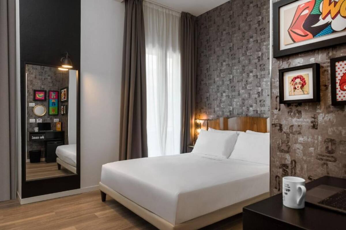 A hotel room with a bed, a nightstand, a lamp, a coffee mug, and artwork on the wall.