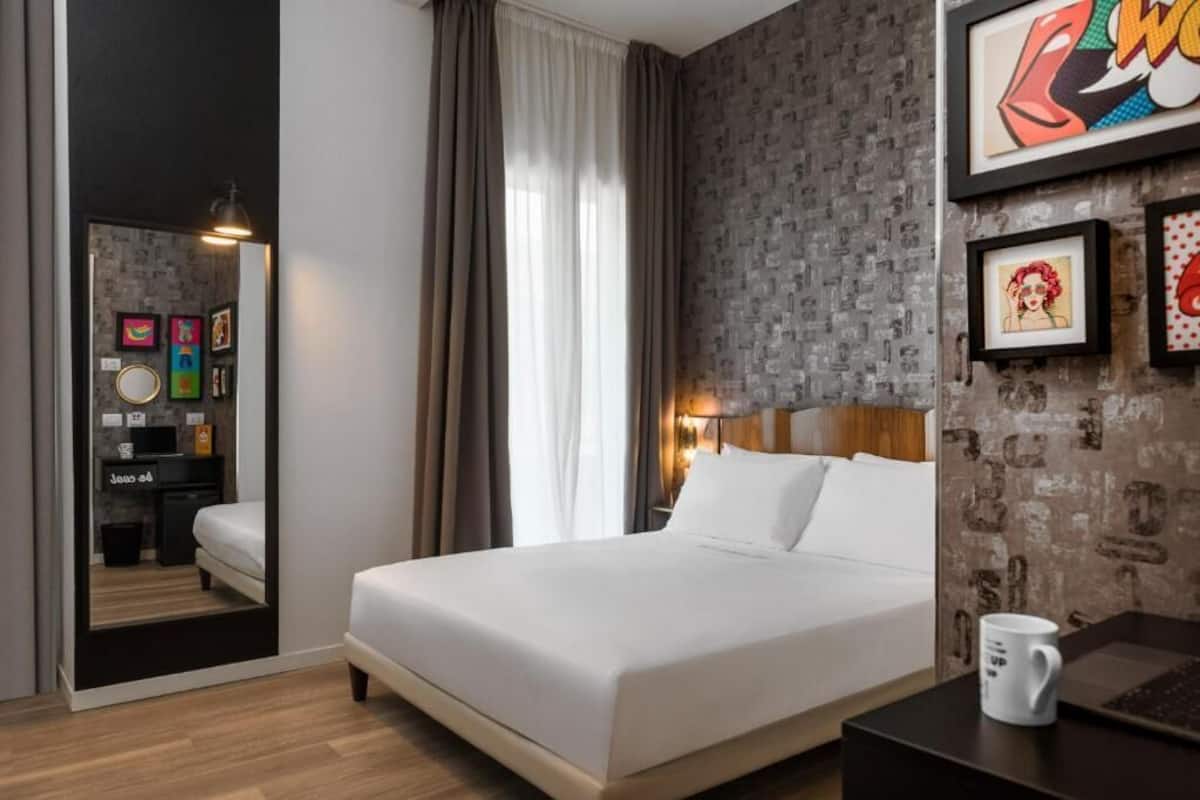 A hotel room with a bed, a nightstand, a lamp, a coffee mug, and artwork on the wall.