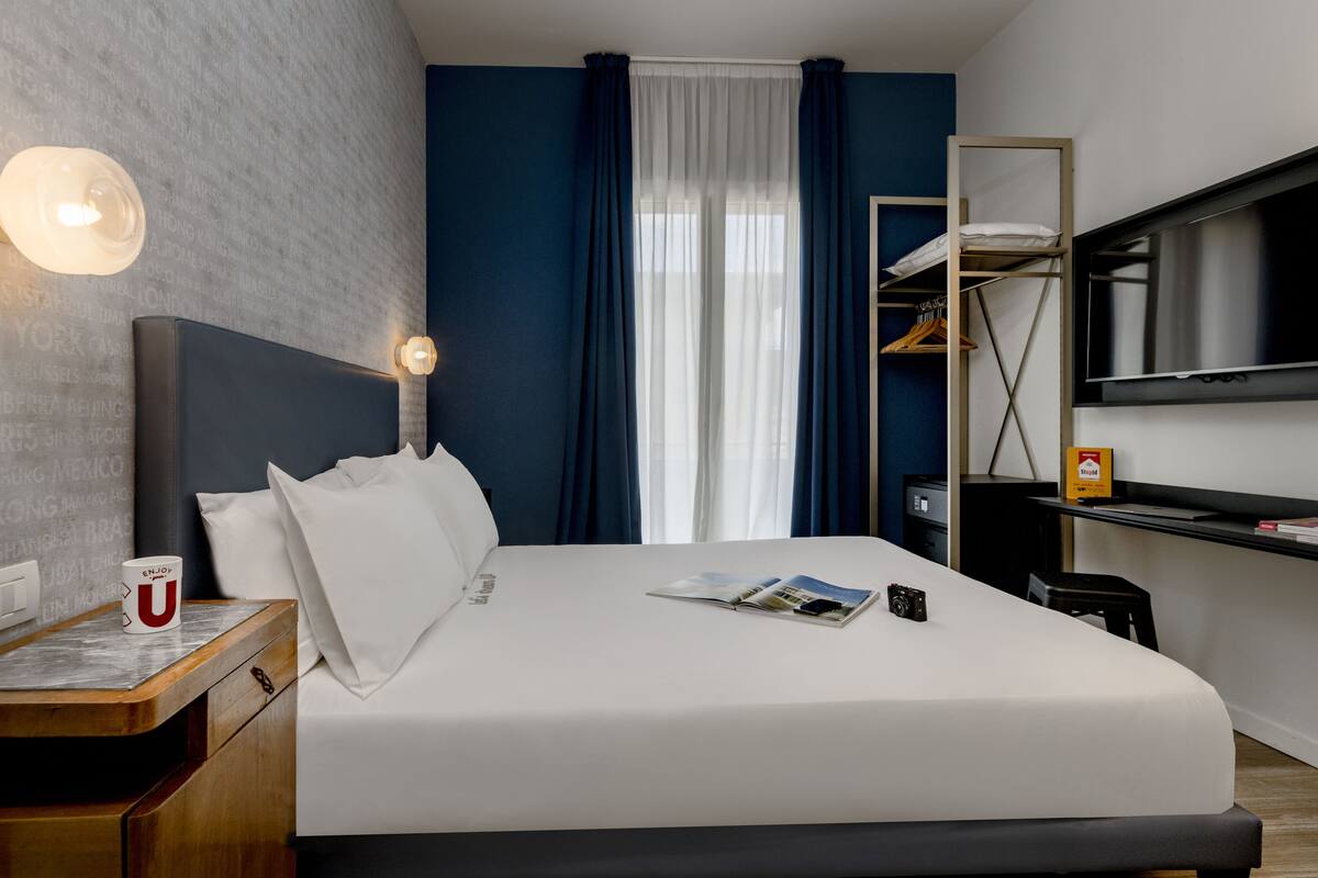 A modern hotel room with a bed, a bedside table with a cup, a magazine, and a camera. There is a TV on the wall and a wardrobe with hanging clothes.