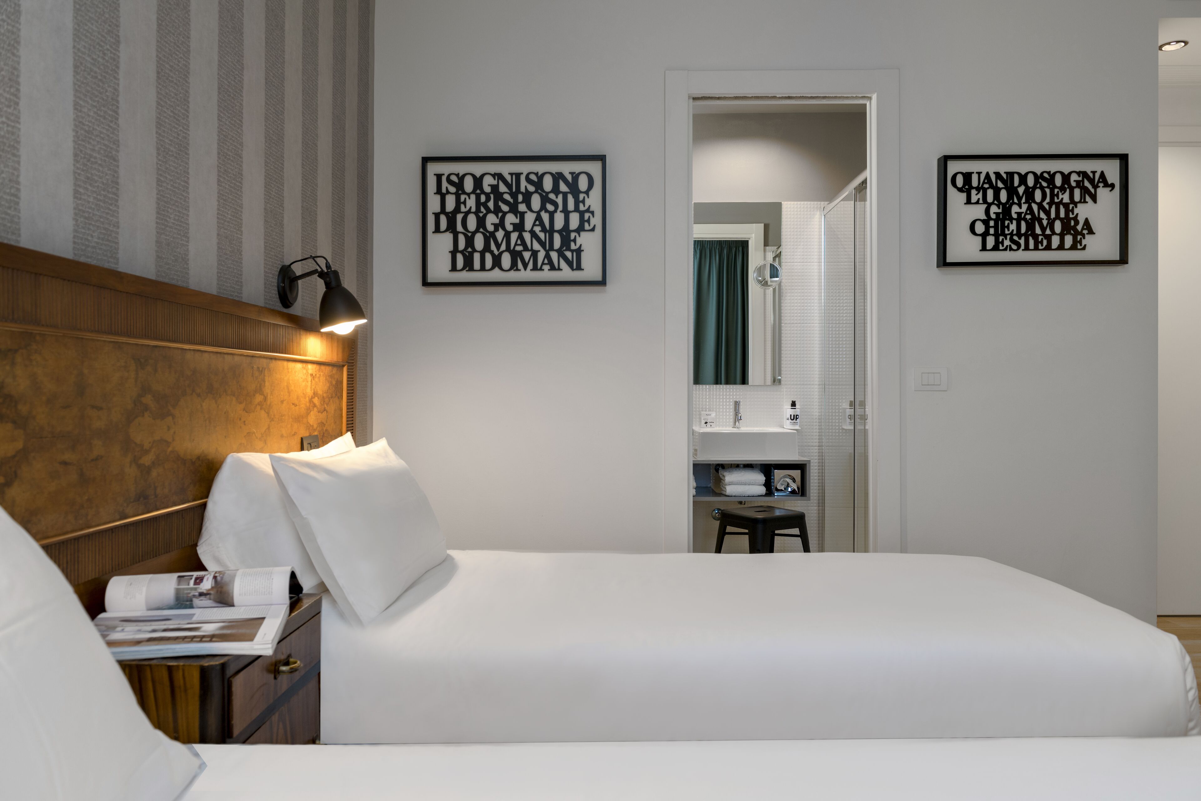 A hotel room with a large bed, a wooden headboard, a nightstand with a lamp, and two framed quotes on the wall.