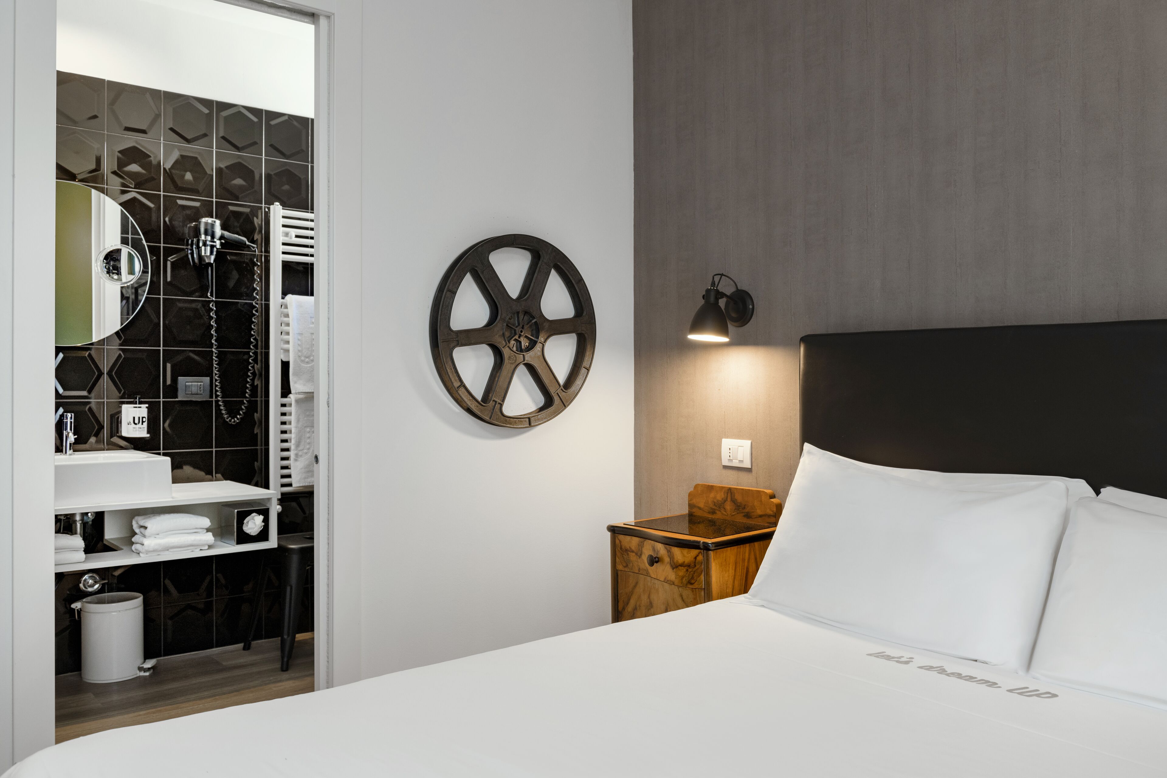 Ein Hotelzimmer mit einem Bett, einem Nachttisch aus Holz, einer Wandlampe, einem runden Spiegel und einem Badezimmer mit Handtuchhalter und Waschbecken.