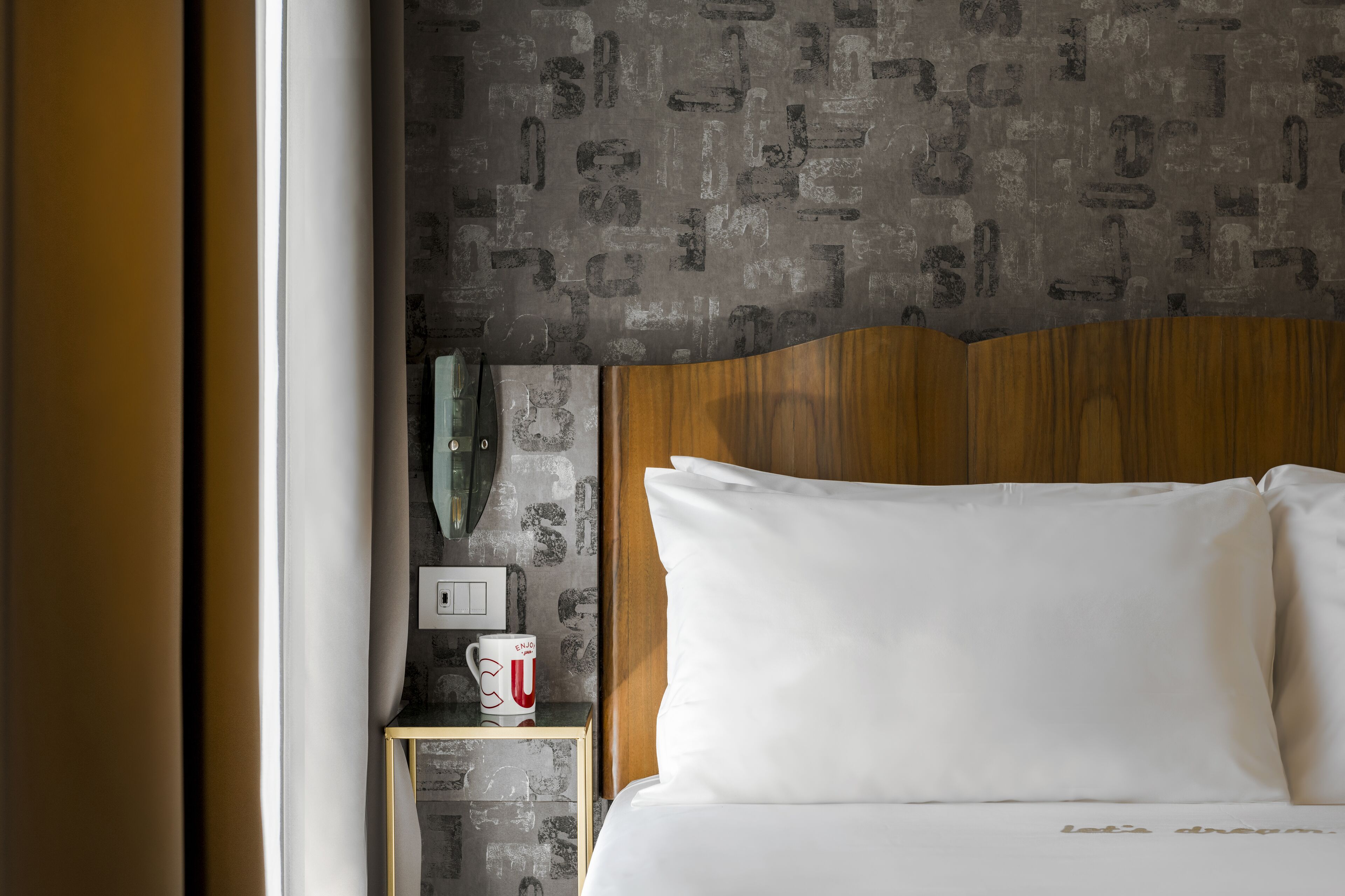 A hotel room with a wooden headboard, a white bed, a nightstand with a white mug, and a textured wall.