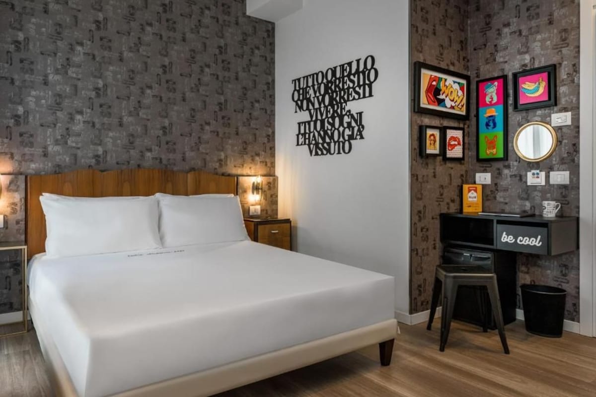 A modern bedroom with a large bed, a desk, and a wall with framed artwork.