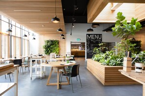 Una cafetería moderna con mesas y sillas de madera, un panel de menú y plantas de interior.