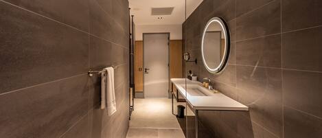 Une salle de bain moderne avec des murs recouverts de carreaux foncés, un lavabo blanc et un miroir doté d’un cadre circulaire.