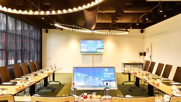A conference room with a long table, chairs, a television screen displaying 'Stenden Hotel', and a window with curtains.