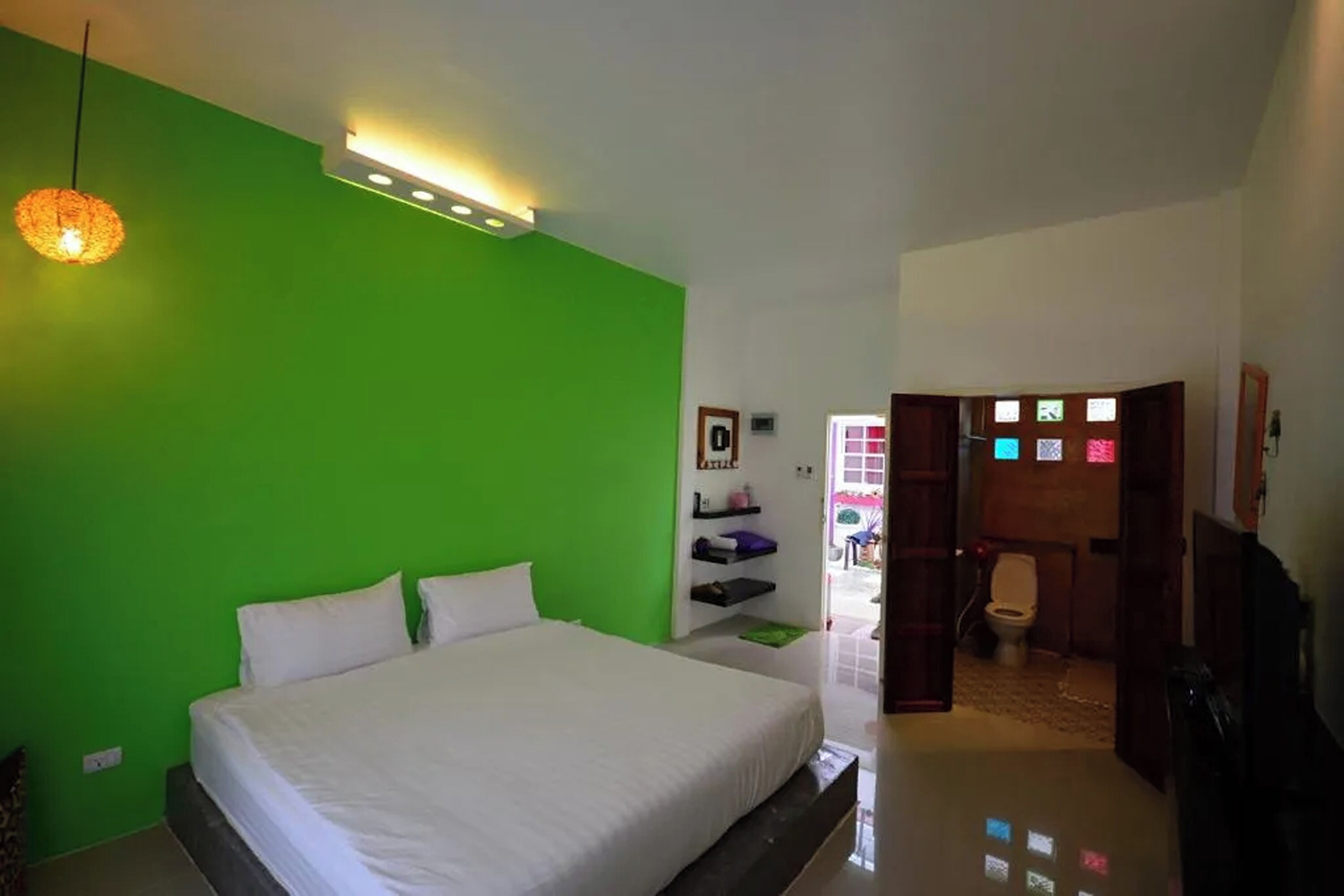 A bedroom with a green wall, a bed with white bedding, a hanging light fixture, and a door leading to a bathroom.