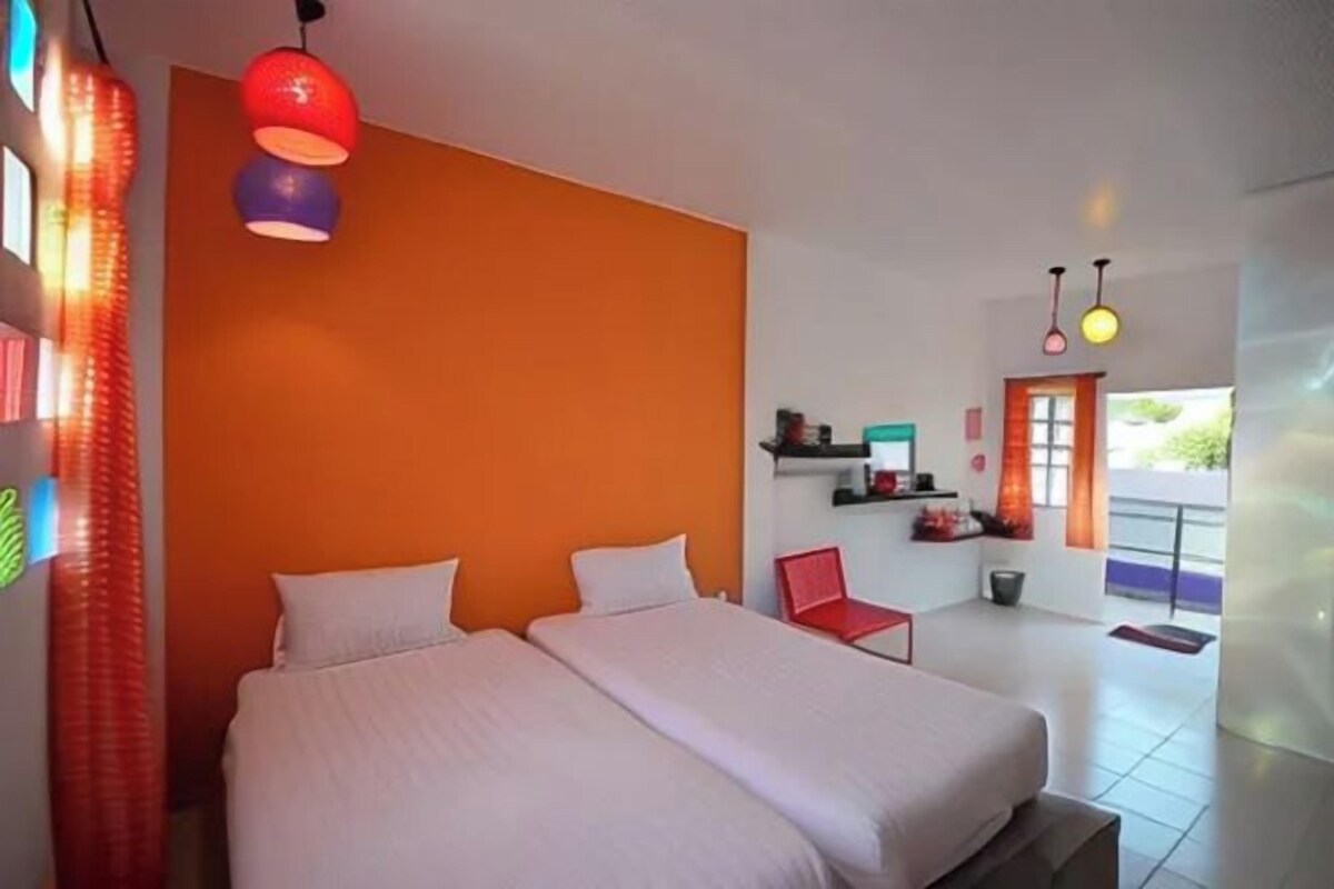 Une chambre avec deux lits, un mur d’accent orange et une chaise rouge.