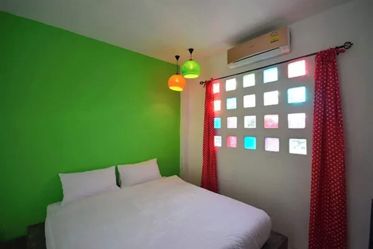 A room with a green wall, a bed with white bedding, a window with colorful square lights, and a red curtain.