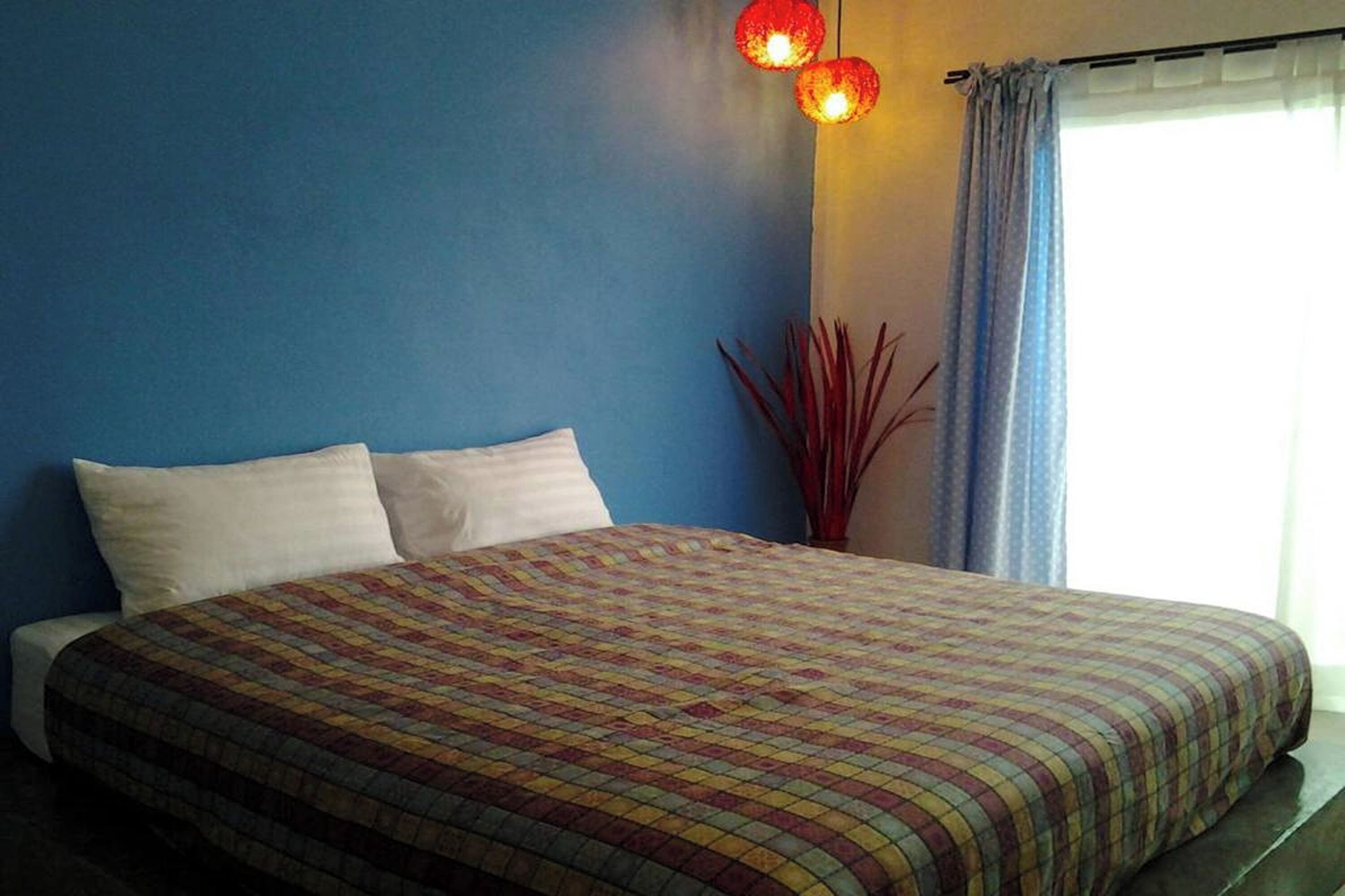 Un dormitorio con cama, pared azul, ventana con cortinas y un accesorio de luz rojo fijado a la pared.
