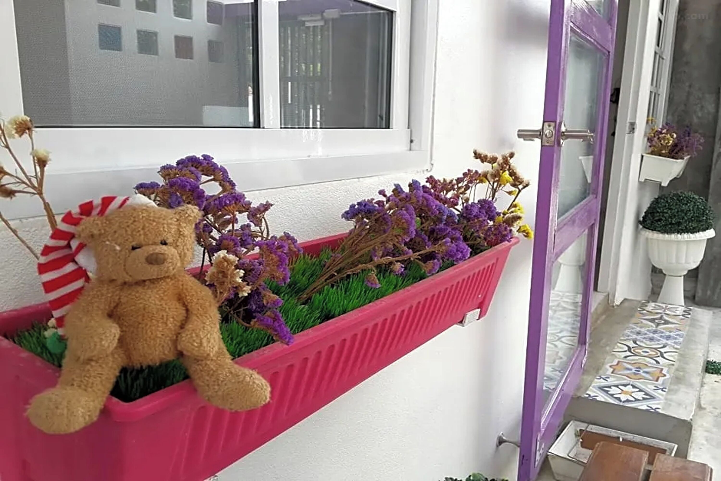 Vensterbank met een roze plantenbak, paarse bloemen en een teddybeer.