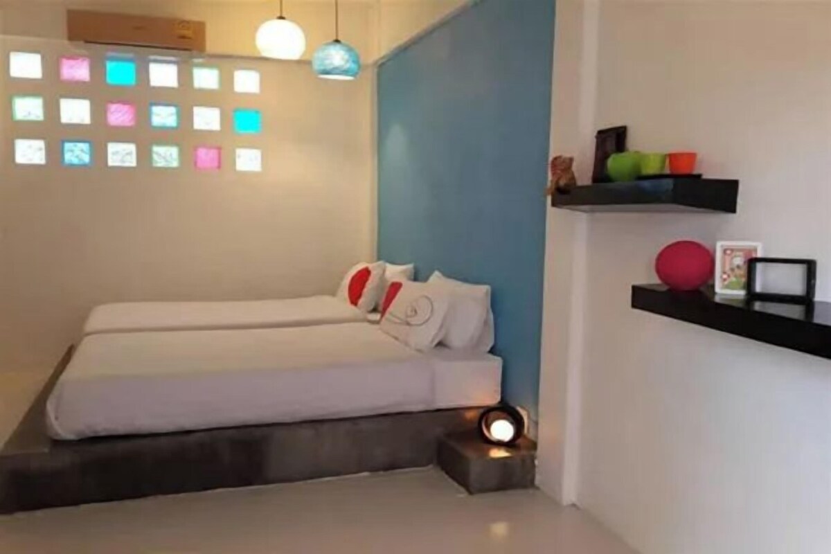 A bedroom with two beds, a blue accent wall, a shelf with decorative items, and a wall-mounted light fixture.