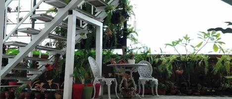 Un espacio de estar al aire libre cubierto con sillas y mesas de metal blanco, rodeado de plantas en macetas y una escalera.