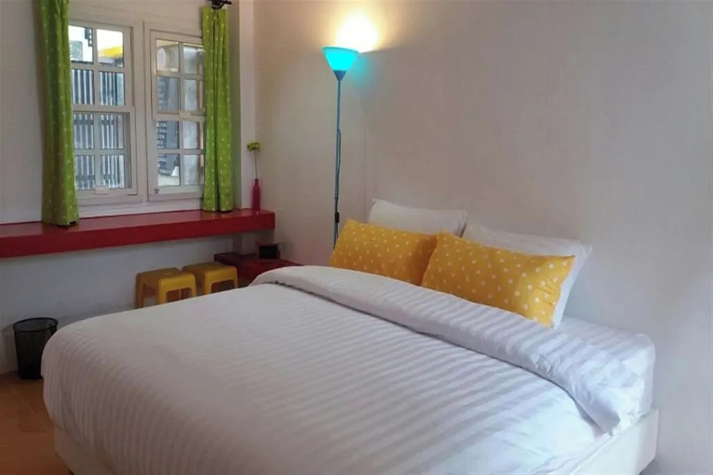 Una cama bien hecha con almohadas blancas y amarillas, una lámpara de pie y una ventana con cortinas verdes.