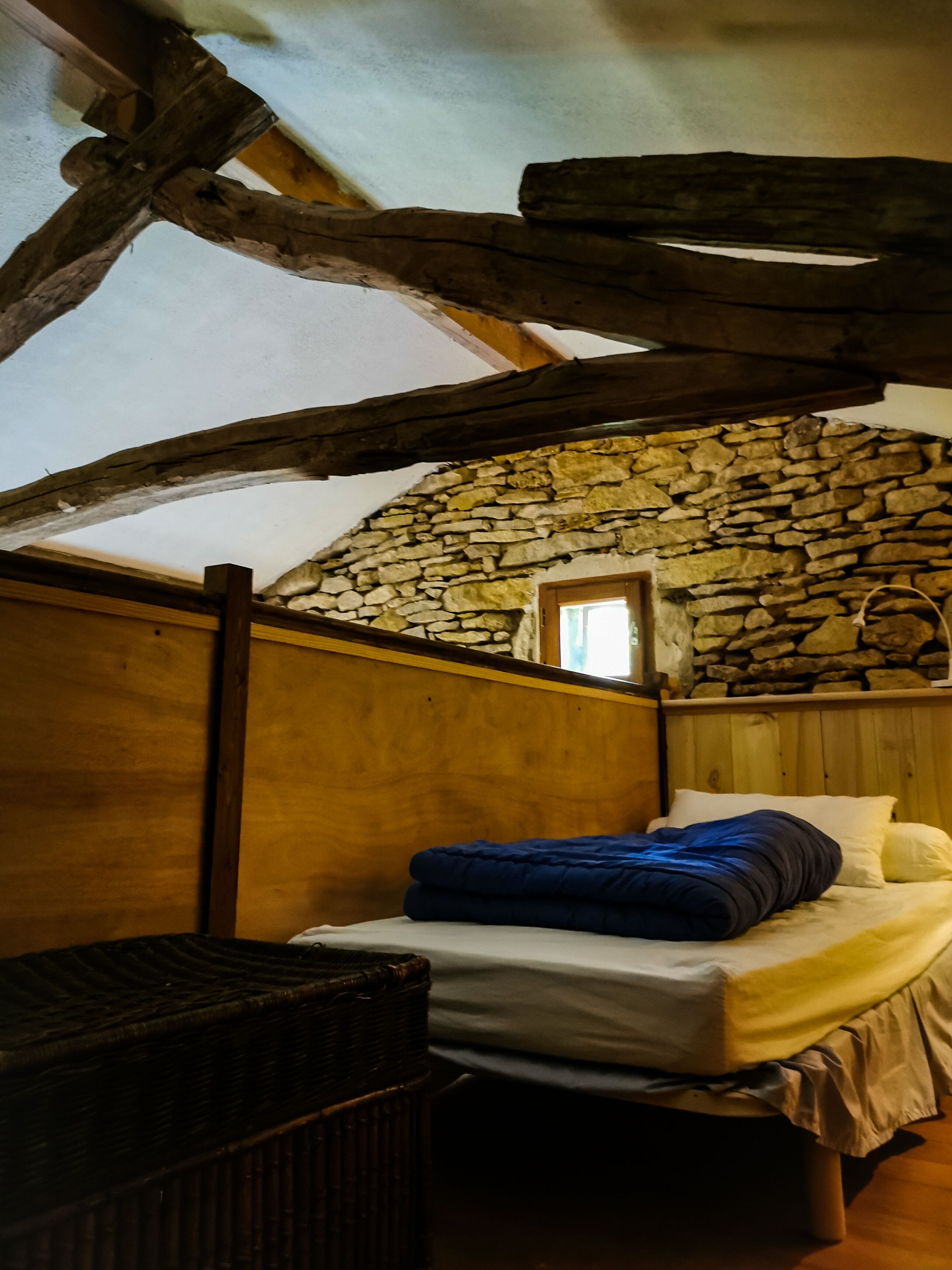 A rustic bedroom with a stone wall, wooden beams, and a bed with a blue blanket.