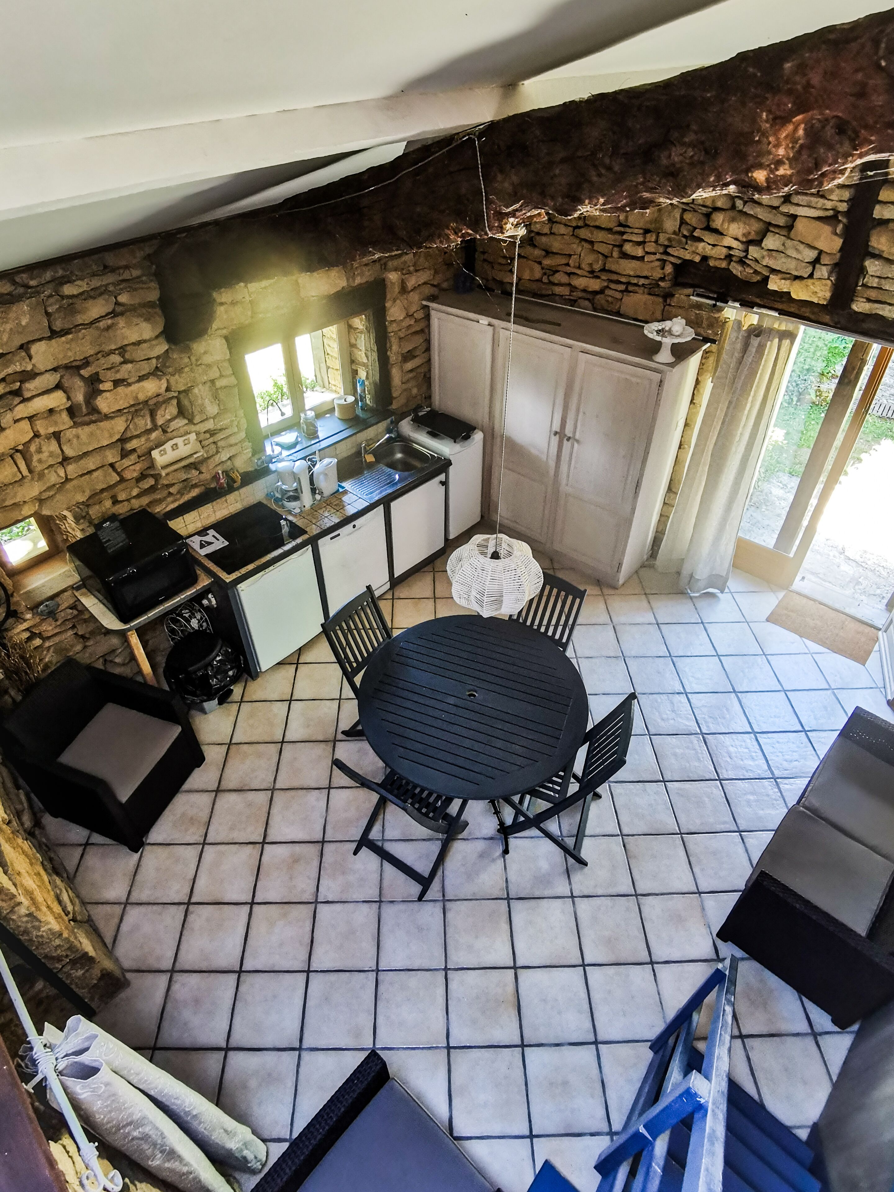 Un espace patio agrémenté d’un mur en pierre, d’une table ronde avec des chaises, d’une petite cuisine équipée d’un évier et d’une cuisinière, et offrant une vue sur la verdure environnante.