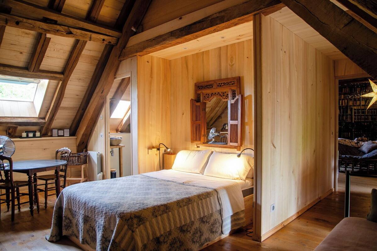 Une chambre mansardée chaleureuse avec un plafond en bois, un lit recouvert d’une courtepointe à motifs, une petite table avec des chaises et une bibliothèque remplie de livres et ornée d’une décoration en forme d’étoile.