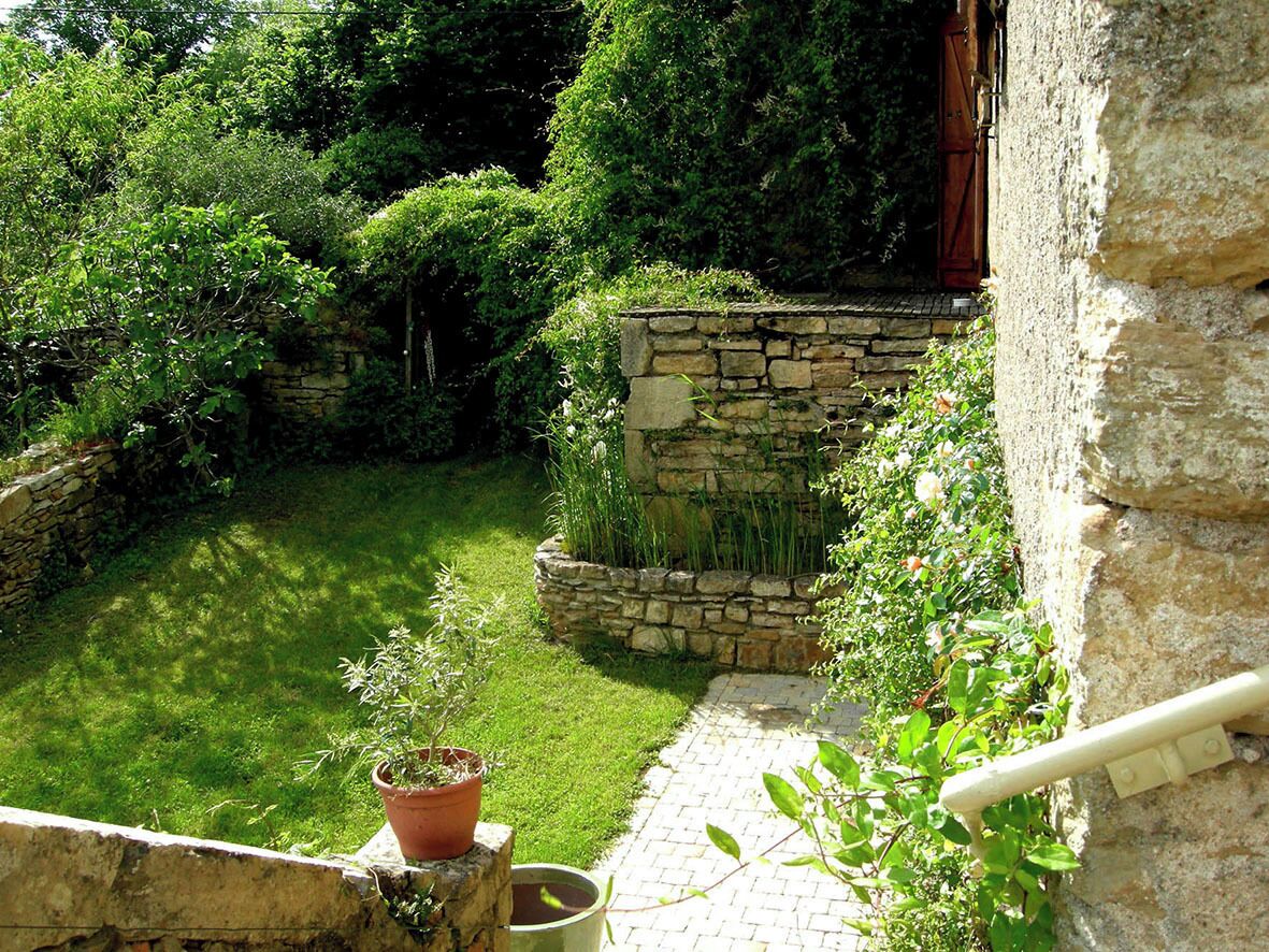 Jardín con muro de piedra, un sendero, planta en maceta y abundante vegetación.