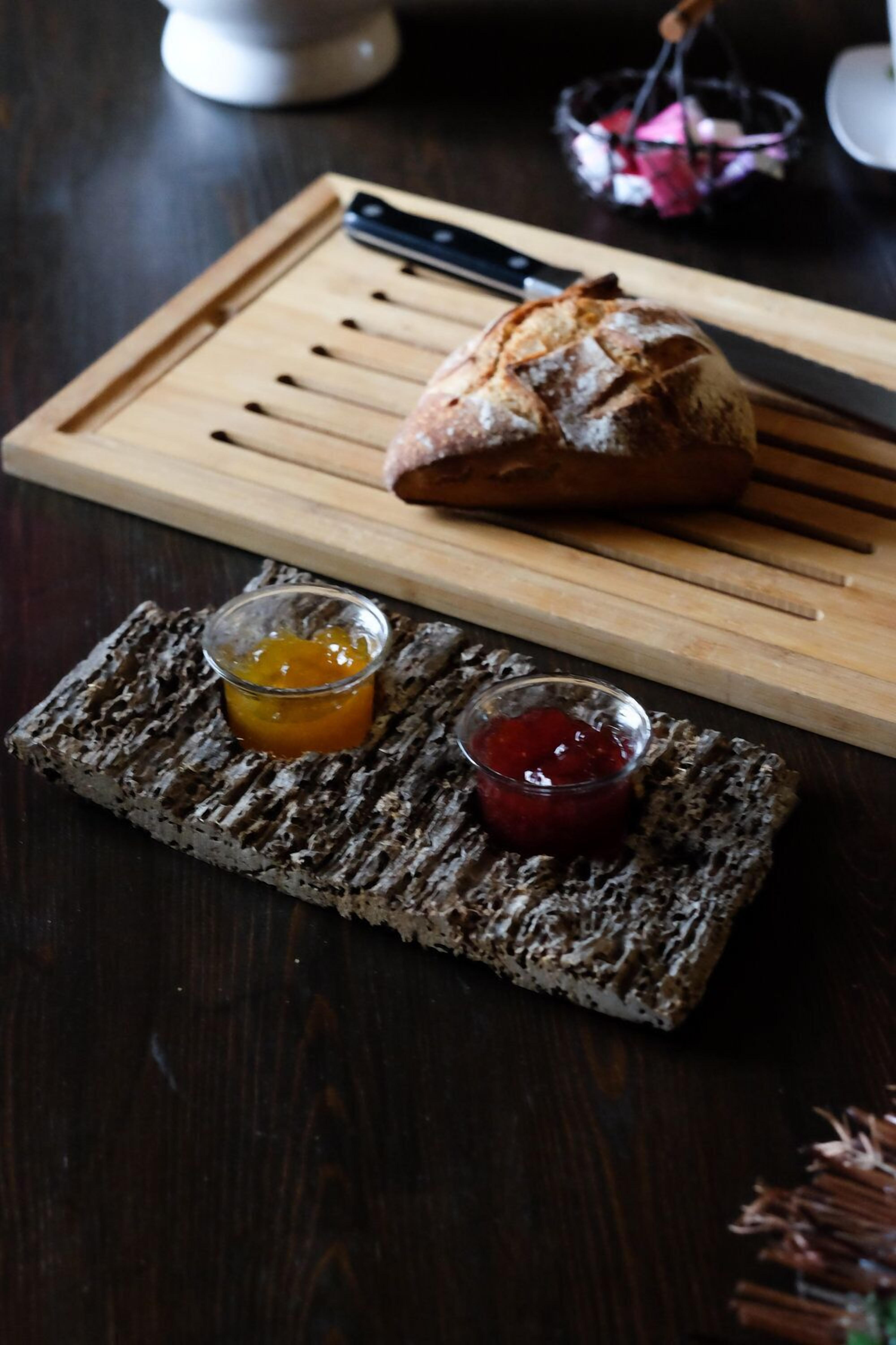 A rustic wooden cutting board with a loaf of bread and two small glass containers of jam.