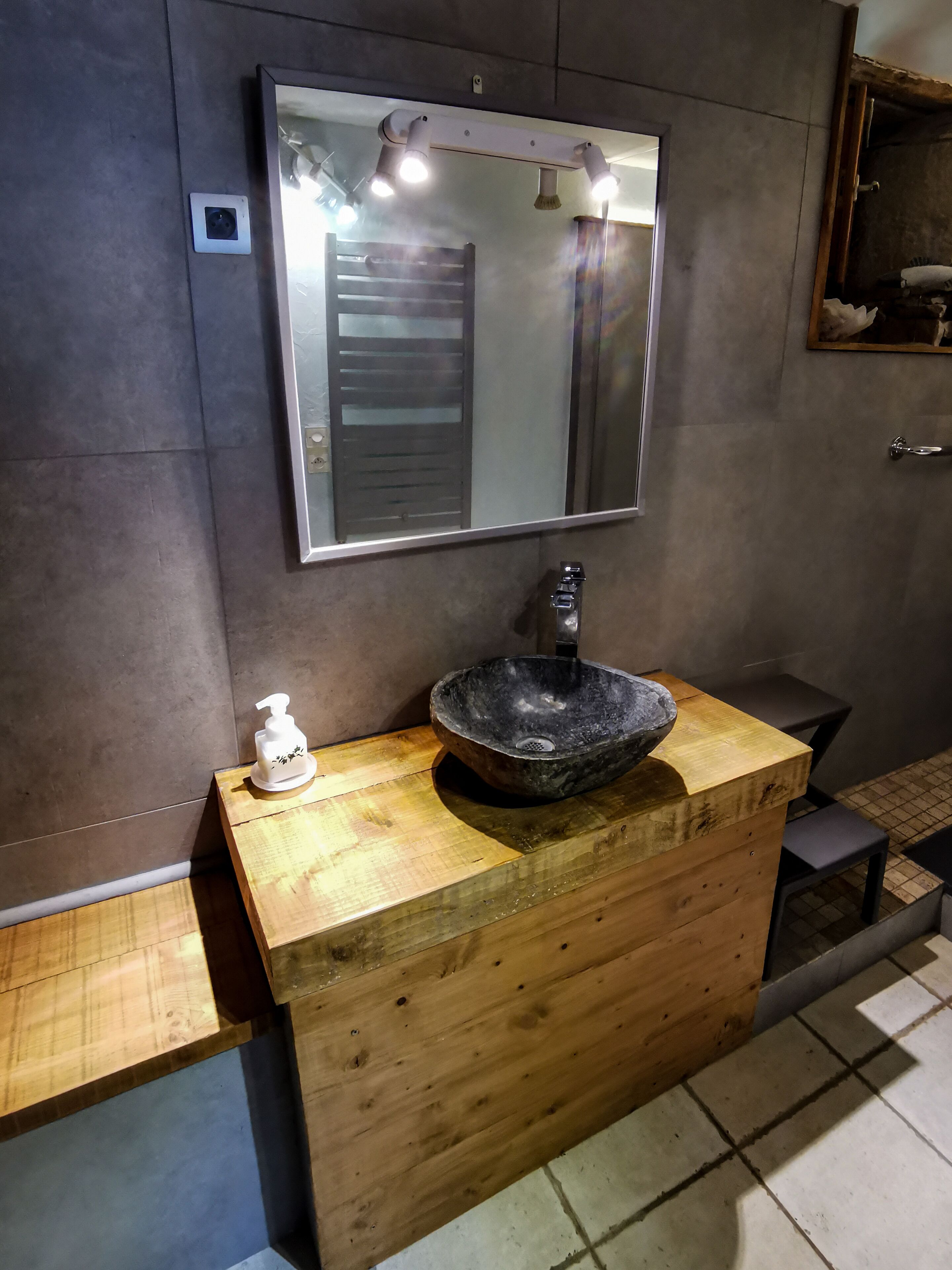Un baño con tocador de madera, lavamanos tipo cuenco negro, espejo y dispensador de jabón.