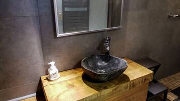 Un baño con tocador de madera, lavamanos tipo cuenco negro, espejo y dispensador de jabón.