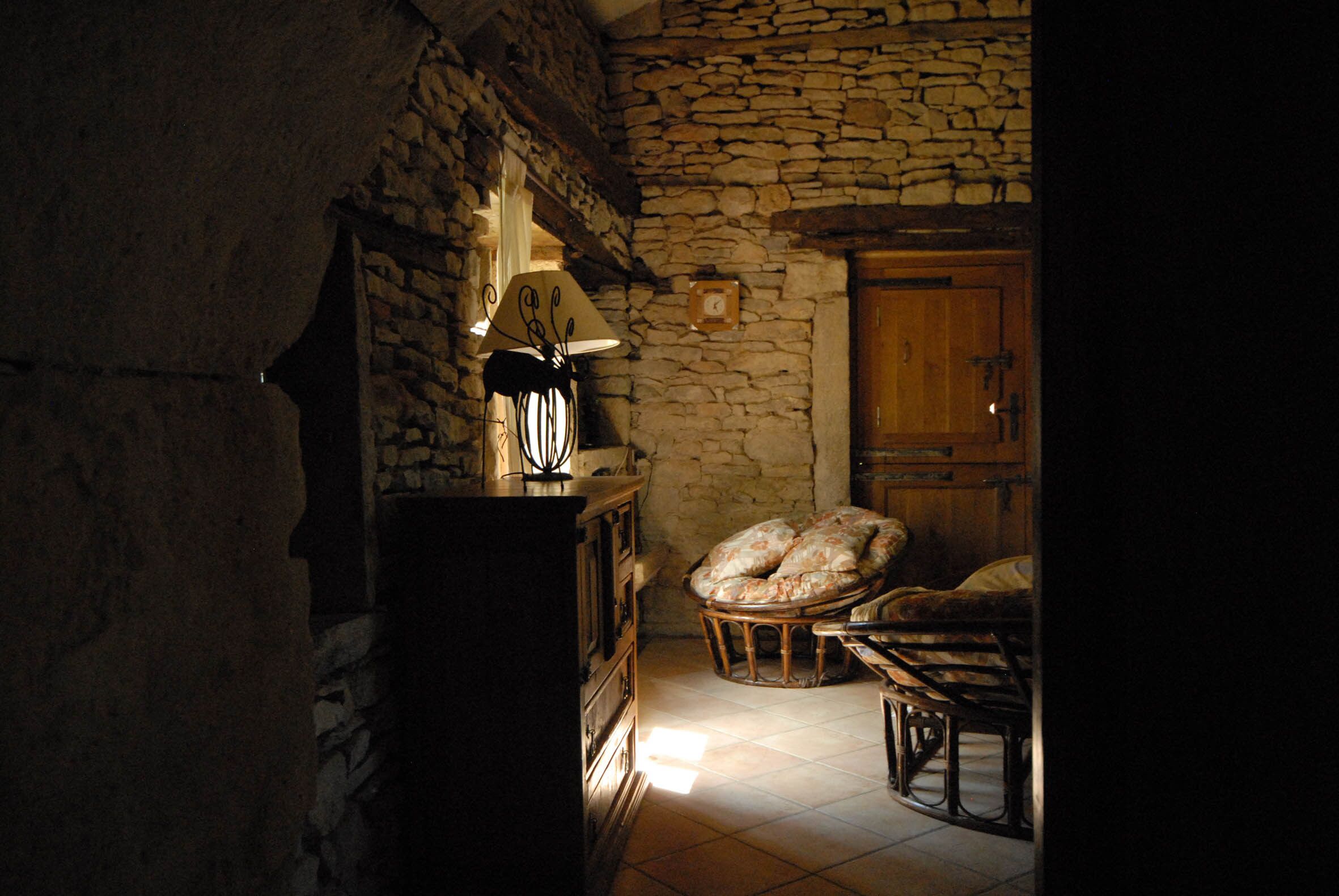 A dimly lit room with stone walls, a wooden cabinet, a lamp, and a chair with cushions.