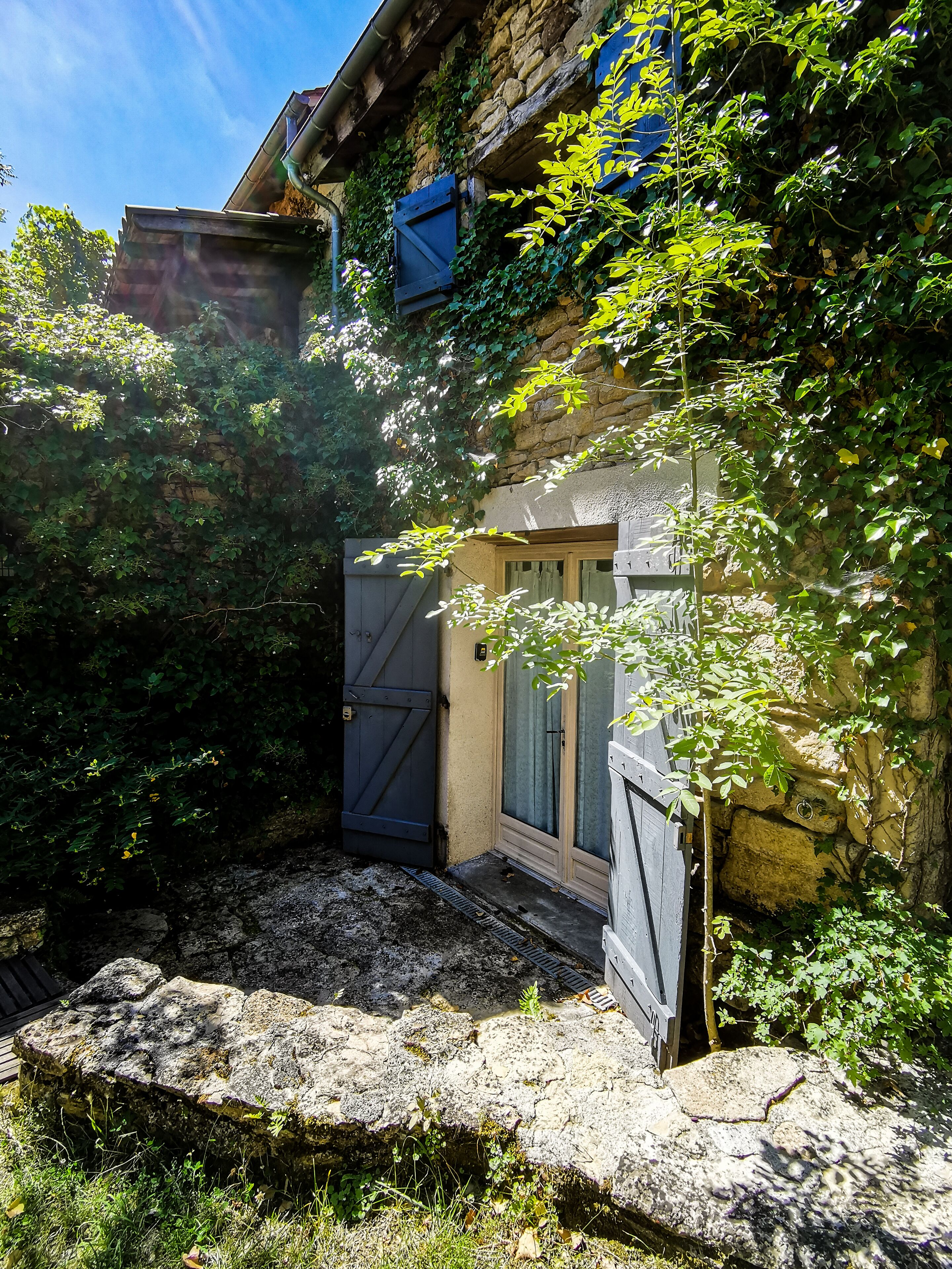 A stone building with a wooden door and shutters, surrounded by greenery and a stone wall.